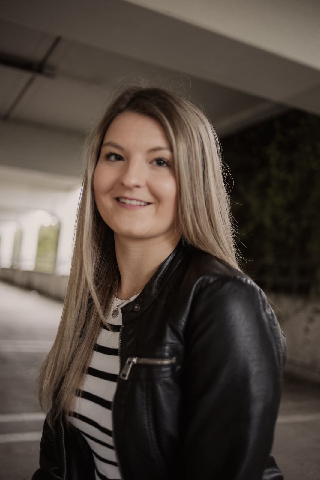 Eine junge Frau mit langen Haaren und einem freundlichen Lächeln trägt eine Lederjacke und ein gestreiftes Shirt. Der urbane Hintergrund verleiht dem Bild eine moderne Atmosphäre.