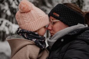 eine Mutter und Ihr Kind berühren liebevoll ihre Nasen im verschneiten Wald, Beide tragen warme Mützen und Schals, ein emotionaler inniger Moment voller Nähe