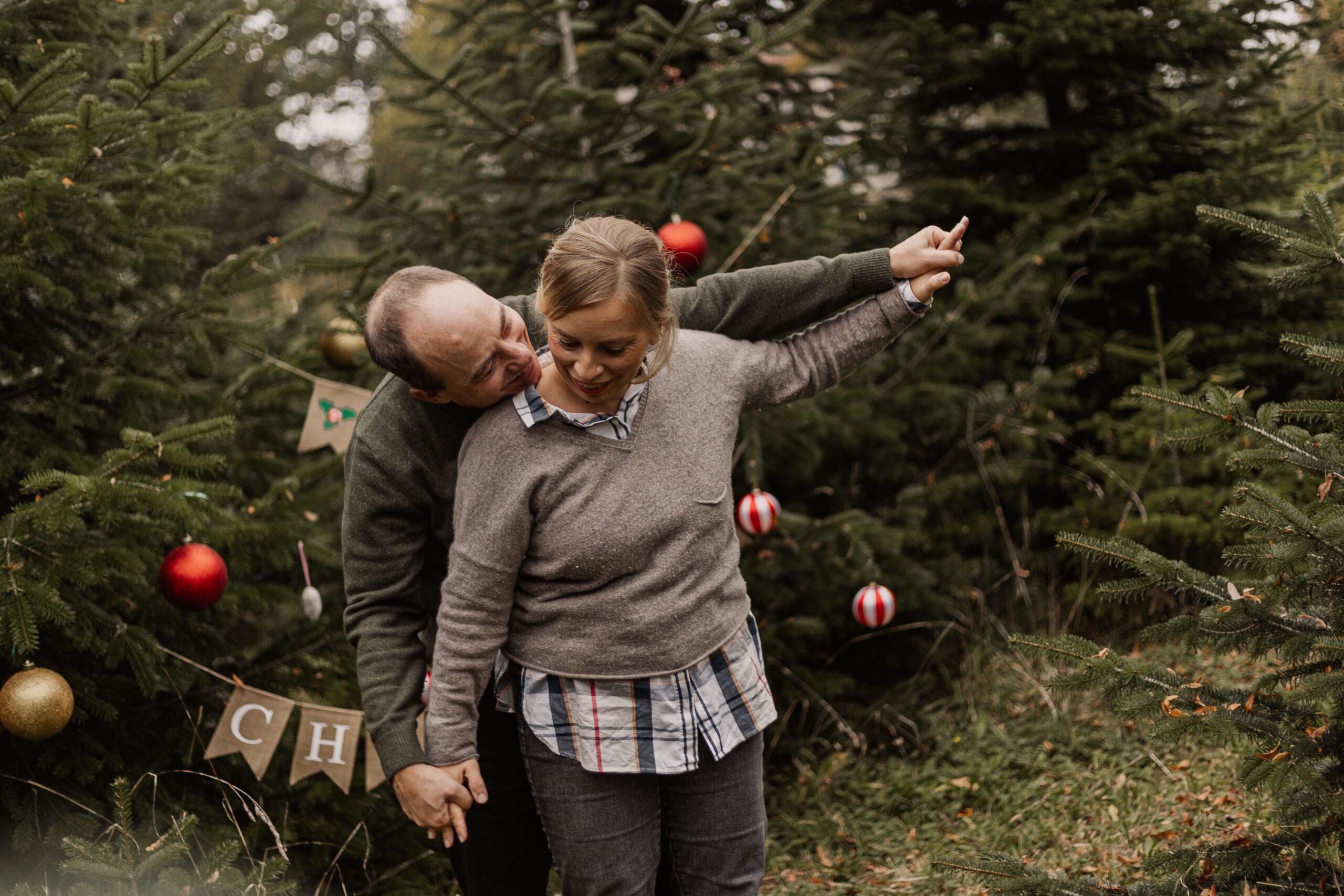 Ein paar sitzt bei einen winterlichen Outdoor Weihnachtsshooting auf einer Decke zwischen kleinen Tannenbäumen, lacht sich an und genießt die gemeinsame Zeit in der Natur