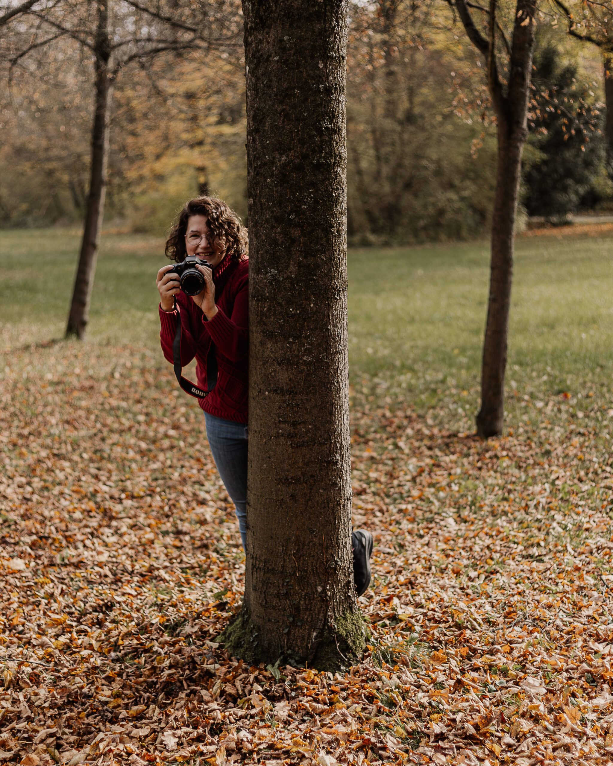 Familienfotografin Sandra von Matelis Fotografie schaut mit Kamera hinter einem Baum hervor im herbstlichen Park im Landkreis Ansbach